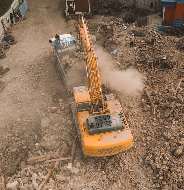 An orange hydraulic excavator with the branding 'HEAVY’S U' is actively engaged in a construction or demolition site, positioned centrally on a ground covered with a mixture of dirt, rubble, and broke