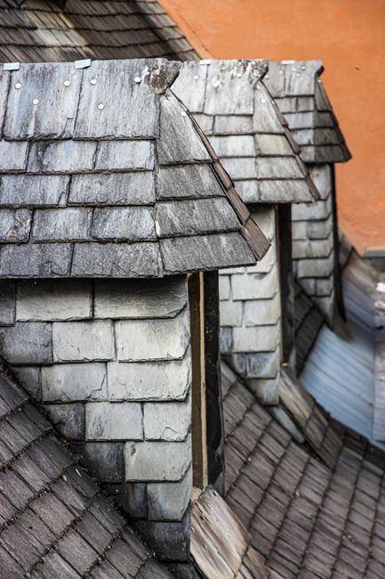 The image depicts a close-up view of a weathered chimney on a sloped roof, covered with gray, overlapping wooden shingles that show signs of age and slight deterioration. The shingles are textured wit