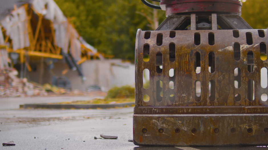 A close-up view of a large, rusted metal demolition weight with multiple rectangular cutouts, positioned on a concrete surface. The background features blurred construction equipment, including yellow